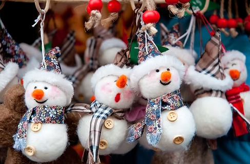 Handmade felt snowman decorations with colourful hats and scarves hanging from a festive market display, surrounded by other Christmas ornaments and red bead garlands.