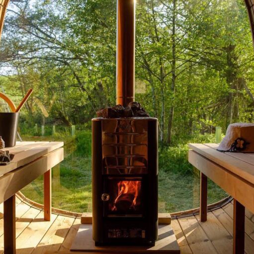 Wood-fired sauna interior with a glowing stove and wooden benches, overlooking a sunlit woodland landscape through large panoramic windows.