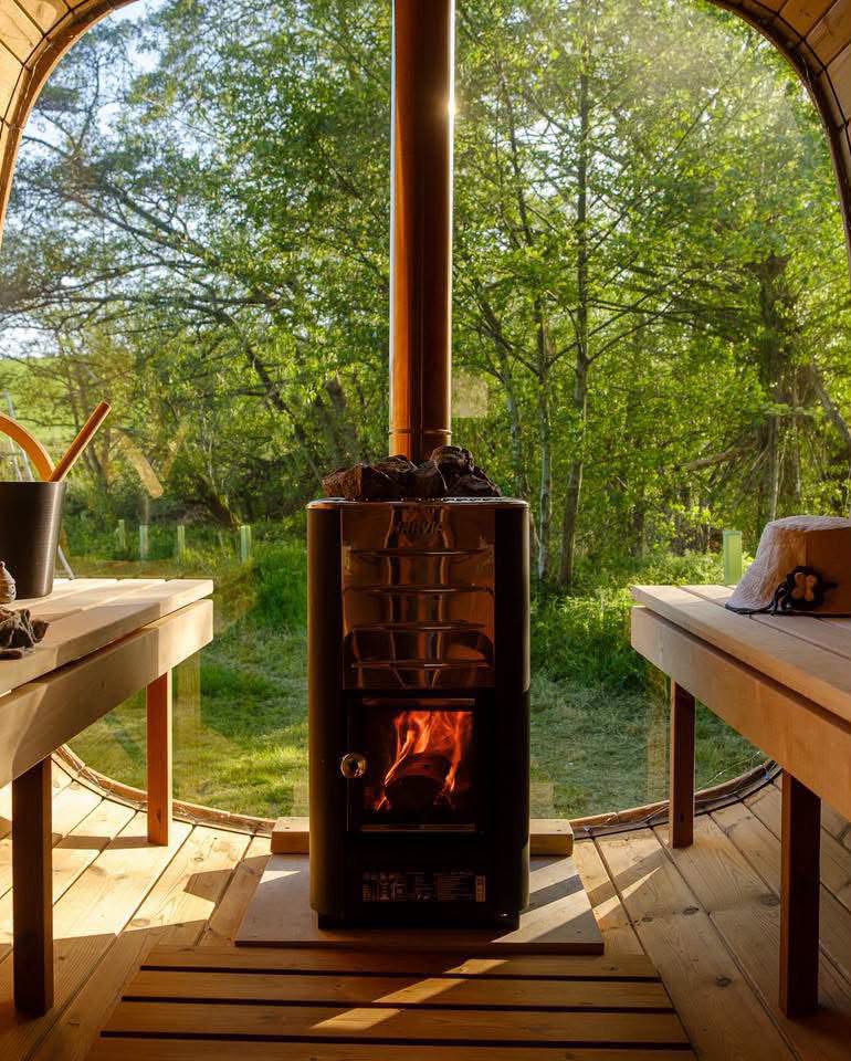 Wood-fired sauna interior with a glowing stove and wooden benches, overlooking a sunlit woodland landscape through large panoramic windows.