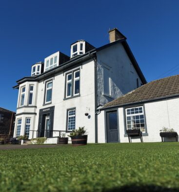 Exterior view of a three-story white building labeled "Seaview" with dark grey trim, dormer windows, and a neat artificial lawn in the foreground under a clear blue sky.