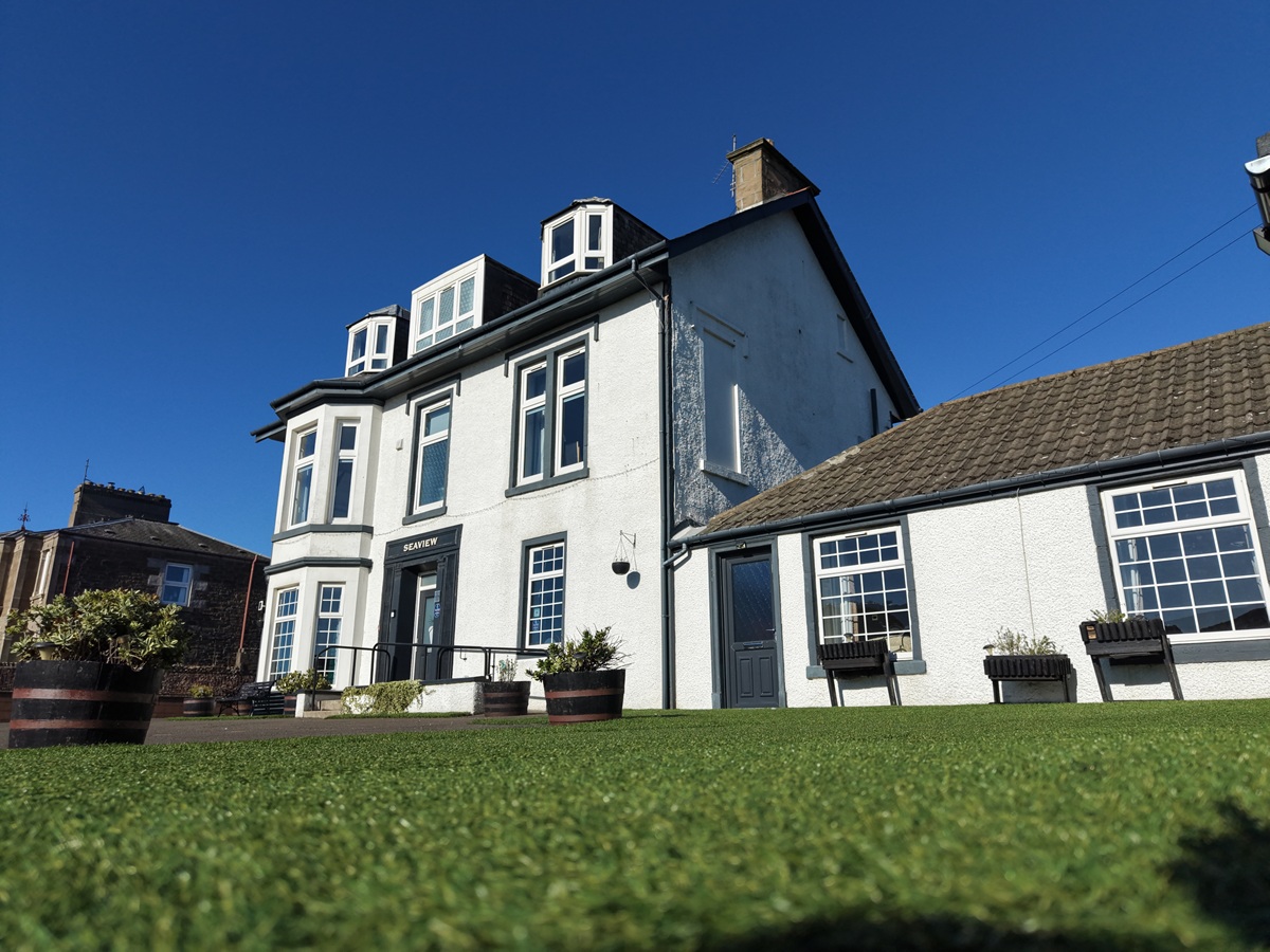 Exterior view of a three-story white building labeled "Seaview" with dark grey trim, dormer windows, and a neat artificial lawn in the foreground under a clear blue sky.