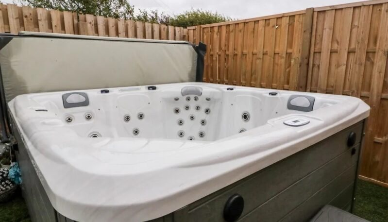 Outdoor hot tub with its lid open, set on artificial grass enclosed by a wooden privacy fence.