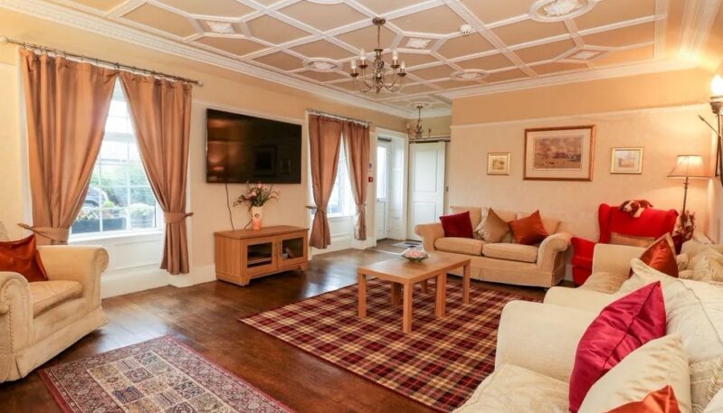 Spacious living room with an ornate coffered ceiling, multiple beige sofas accented with red cushions, hardwood floors, and a red plaid area rug.