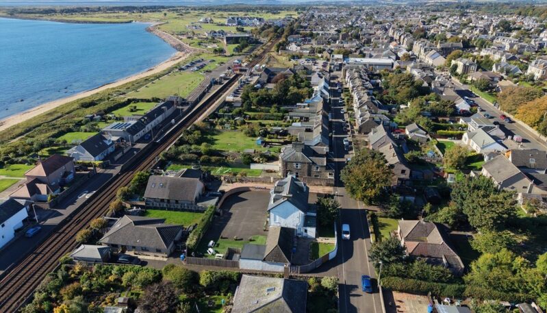 High-angle aerial view of a coastal town, showing a railway line running parallel to a sandy beach and blue sea on the left, alongside residential streets and buildings stretching inland.