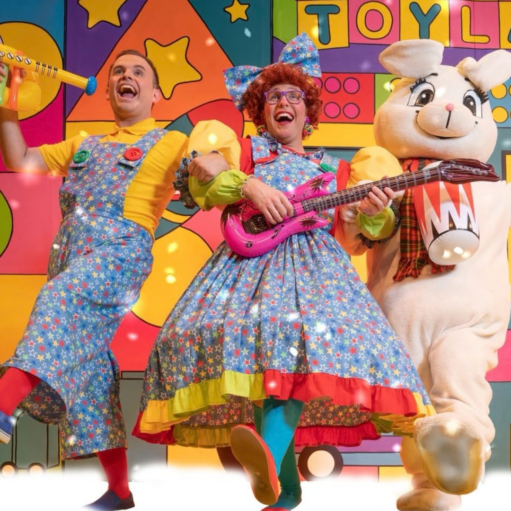 Colourful promotional image for a family pantomime featuring two performers in bright star-patterned costumes and a large rabbit character, standing in front of a vibrant toy-themed backdrop with oversized shapes and the word “TOYLAND”.