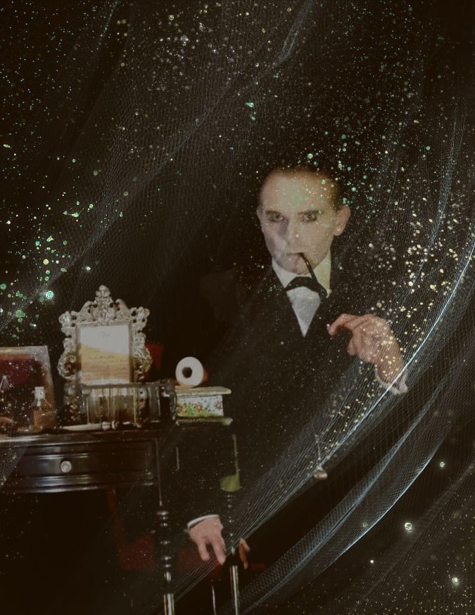 Atmospheric promotional image showing a performer in formal evening attire standing beside a dressing table, partially obscured by sparkling veil-like fabric. The dark, vintage-style scene features theatrical lighting, decorative props and a mysterious cabaret-inspired aesthetic.