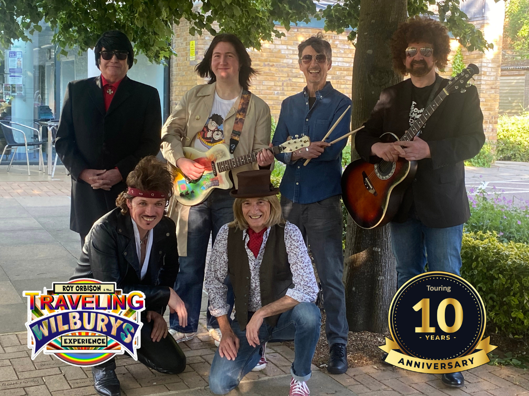 Promotional image for the Roy Orbison and the Traveling Wilburys Experience, featuring six musicians posing outdoors with guitars and performance props beneath a tree. The image includes the show logo and a gold badge highlighting a 10-year anniversary tour.