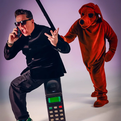 Promotional image featuring a performer in dark clothing and sunglasses seated on an oversized vintage mobile phone while speaking into a handset, with a person in a large red dog costume standing behind. The studio-style image has a simple purple background and a playful, surreal comedy theme.