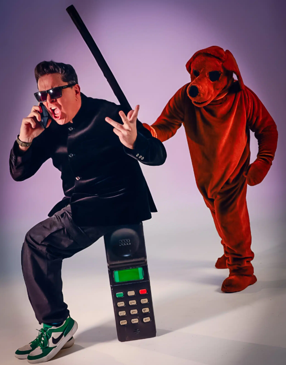 Promotional image featuring a performer in dark clothing and sunglasses seated on an oversized vintage mobile phone while speaking into a handset, with a person in a large red dog costume standing behind. The studio-style image has a simple purple background and a playful, surreal comedy theme.