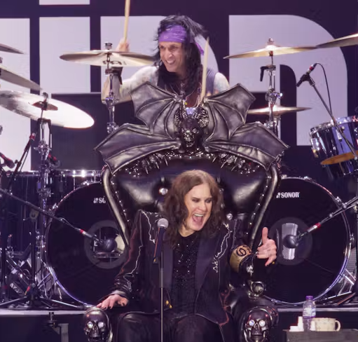 Promotional concert image featuring a vocalist seated in an ornate black throne at the front of a stage, with a drummer and drum kit behind. The stage is lit with purple and white lighting, creating a dramatic heavy-rock performance atmosphere.