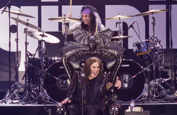 Promotional concert image featuring a vocalist seated in an ornate black throne at the front of a stage, with a drummer and drum kit behind. The stage is lit with purple and white lighting, creating a dramatic heavy-rock performance atmosphere.