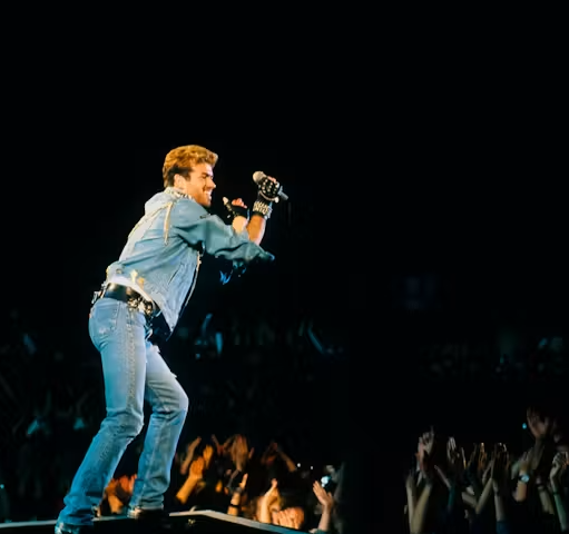 Performer singing into a microphone while standing on a concert stage above a crowd, wearing a denim outfit and gloves. Audience members can be seen raising their hands beneath the spotlight-lit stage.