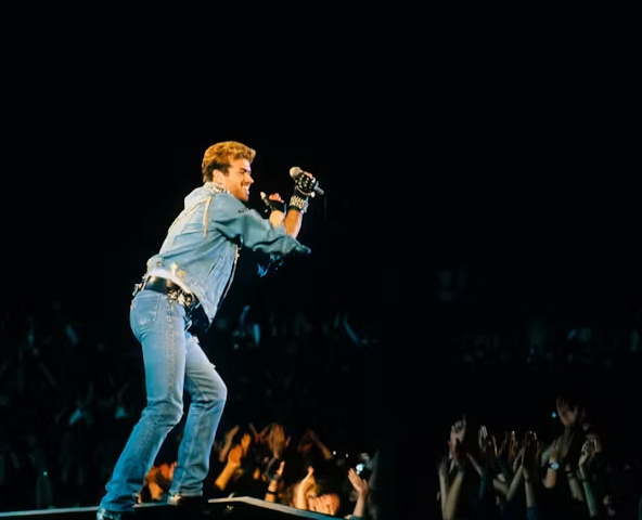 Performer singing into a microphone while standing on a concert stage above a crowd, wearing a denim outfit and gloves. Audience members can be seen raising their hands beneath the spotlight-lit stage.