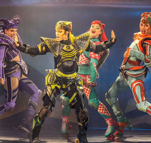 Promotional image for Starlight Express featuring performers in brightly coloured futuristic costumes and roller skates. The performers are posed mid-performance under theatrical stage lighting, wearing detailed armour-inspired outfits in purple, yellow, turquoise and red.