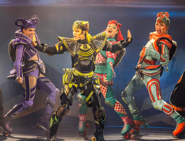 Promotional image for Starlight Express featuring performers in brightly coloured futuristic costumes and roller skates. The performers are posed mid-performance under theatrical stage lighting, wearing detailed armour-inspired outfits in purple, yellow, turquoise and red.