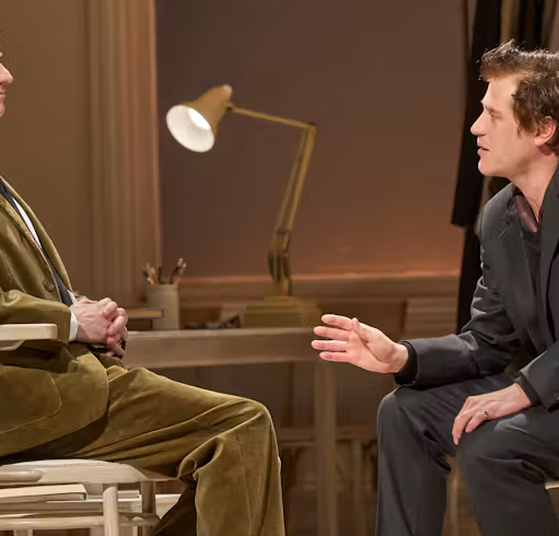 Stage production image showing two performers seated facing each other in a warmly lit room. One performer sits upright in a chair while the other leans forward in conversation, with a desk lamp and simple theatre set visible in the background.